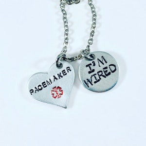 May include: A silver-colored necklace with two pendants. One pendant is heart-shaped and says "PACEMAKER" with a red medical symbol. The other pendant is round and says "I'M WIRED" in black letters. The necklace chain is also silver.