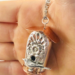 May include: Silver birdhouse pendant necklace with a copper roof and base. The birdhouse features floral detailing and a small silver bird perched on the front. The necklace chain is silver.
