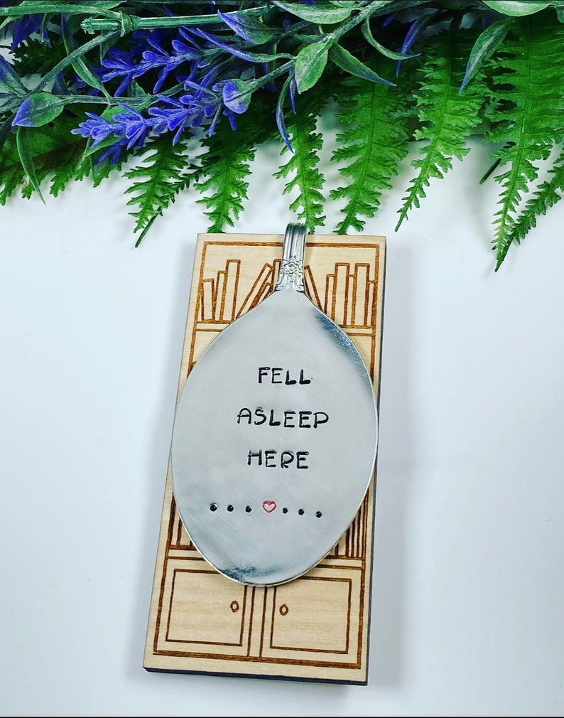 May include: A silver spoon with the words "FELL ASLEEP HERE" engraved on it. The spoon is hanging on a wooden plaque with a design of bookshelves.