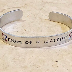May include: Silver bracelet with the text "Mom of a Warrior" and two red, white, and blue ribbon designs.