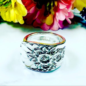 May include: A silver ring made from a repurposed spoon with a floral design. The ring is wide and has a detailed floral pattern.