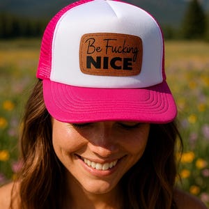May include: A trucker hat with a white front panel and a pink mesh back and brim. The hat features a brown leather patch with the words "Be Fucking NICE" in black lettering. The hat is worn outdoors.