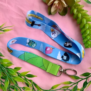 May include: Two blue lanyards with cartoon character designs. One lanyard features a green and lime green pattern, and the other has a light blue background with cartoon characters and white clouds. Both lanyards have a silver clasp.