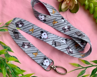 May include: A gray lanyard with a white striped pattern featuring cartoon raccoons and opossums with bananas and trash cans. The lanyard has a silver metal clip.
