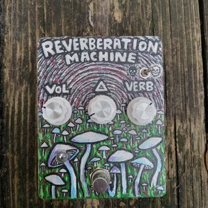 Può includere: Un pedale per effetti per chitarra dipinto su misura con uno sfondo verde, blu e viola con un design a forma di fungo. Il pedale è etichettato "REVERBERATION MACHINE" e ha tre manopole etichettate "VOL", "▲" e "VERB".