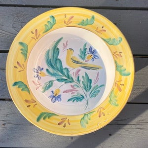 May include: A yellow ceramic bowl with a hand-painted design of a yellow bird perched on a green leafy branch with pink and blue flowers. The bowl has a yellow rim with a gold accent.
