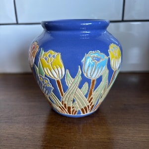 May include: A blue ceramic vase with a floral design featuring yellow, blue, and orange tulips. The vase has a rounded shape and a smooth, glossy finish. The tulips are set against green leaves and brown stems.