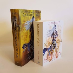 May include: Two hardcover books titled "Kingdom of Ash" by Sarah J. Maas. The larger book has a yellow and brown cover with a warrior figure. The smaller book has a white cover with a warrior figure.