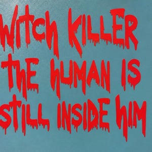 以下が含まれることがあります： 水色の背景に「WITCH KILLER THE HUMAN IS STILL INSIDE HIM」という文字が太く鮮やかな赤色で書かれた正方形の看板。文字は滴り落ちるようなデザインで、濡れたペンキのような質感を表現している。