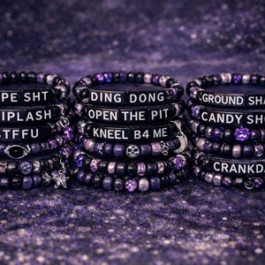 May include: Stacked black and purple beaded bracelets with white lettered phrases like "TYPE SHT" and "WHIPLASH." Each bracelet has unique charms, including stars and skulls. The background is a dark, speckled purple.