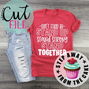 Op de afbeelding: Rood T-shirt met witte tekst die luidt: "Sta niet aan de zijlijn - Sta op Sta sterk Sta samen". Een cupcake met roze glazuur en groene sprinkles bevindt zich in een cirkel met de tekst "Stap weg van de taart" eromheen.