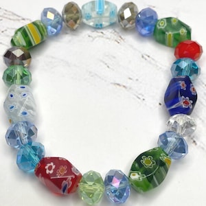May include: A vibrant beaded bracelet with a mix of glass beads in blue, green, red, and clear. The beads are in various shapes, including faceted and patterned designs. The bracelet is set against a white backdrop.