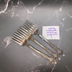 May include: Three silver-plated forks with ornate handles and four tines each. A small white paper note is included with the text: ONEIDA COMMUNITY AFFECTION SILVERPLATE 5. The forks exhibit a metallic sheen with some visible tarnish.