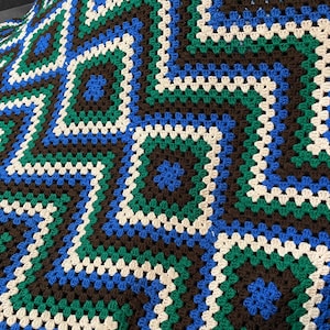 May include: A handmade crochet blanket with a geometric pattern in brown, blue, green, and cream. The design features squares and zig-zag lines, creating a textured look. This cosy blanket is suitable for home decor and adds a touch of handmade charm.