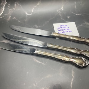 Könnte beinhalten: Drei versilberte Tafelmesser mit einem dekorativen Blumenmuster auf den Griffen. Eine kleine weiße Karte enthält den handgeschriebenen Text: Oneida Community Affection Silver Plate. Die Messer haben polierte Klingen und angelaufene Griffe.