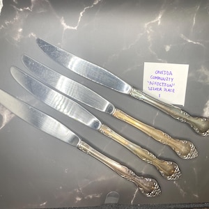 Könnte beinhalten: Vier versilberte Tafelmesser im Oneida Community Affection Muster. Jedes Messer hat eine glatte, polierte Klinge und einen verzierten Griff mit floralen Details. Ein kleiner weißer Zettel trägt die Aufschrift Oneida Community Affection Silver Plate.