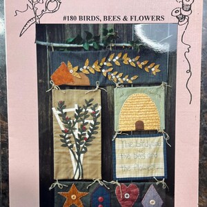 Op de afbeelding: Een handwerkpatroonboekje getiteld Bits & Pieces by Joan #180 Birds, Bees & Flowers. De omslag toont een wandhanger met stoffen panelen die een bijenkorf, een plant op een latwerk en vormen zoals een ster, huis, hart en ruit afbeelden.