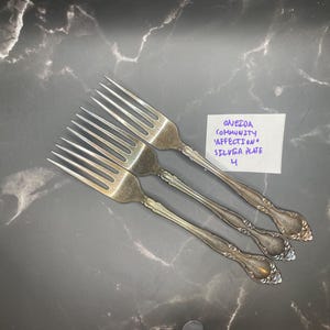 Könnte beinhalten: Drei versilberte Gabeln mit verzierten Griffen, die fächerförmig angeordnet sind. Neben den Gabeln liegt eine kleine weiße Karte mit dem handschriftlichen Text: ONEIDA COMMUNITY 'AFFECTION' SILVER PLATE 4. Die Gabeln haben jeweils vier Zinken.