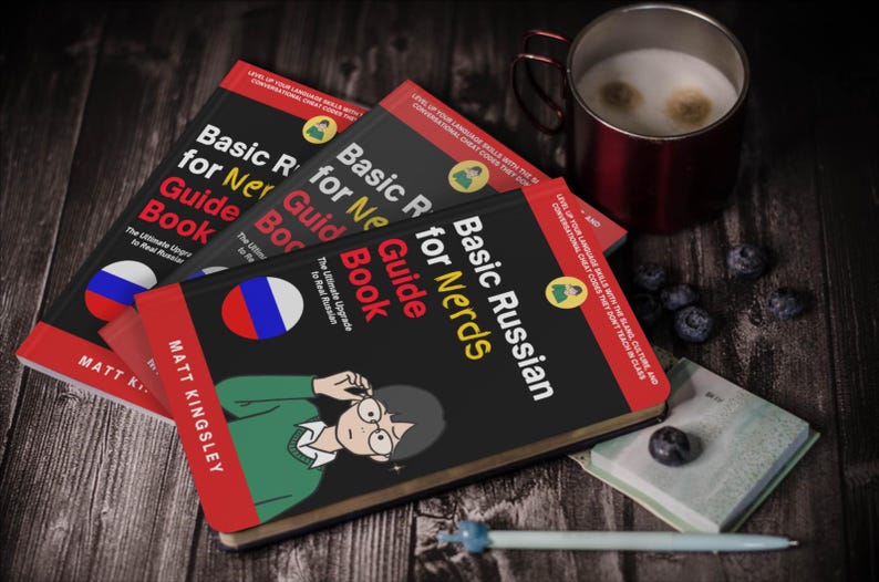 Puede incluir: Tres ejemplares de "Basic Russian for Nerds Guide Book" con una bandera rusa y una ilustraci&oacute;n de dibujos animados. Una taza de caf&eacute;, ar&aacute;ndanos, un bol&iacute;grafo y un peque&ntilde;o cuaderno est&aacute;n sobre una superficie de madera.
