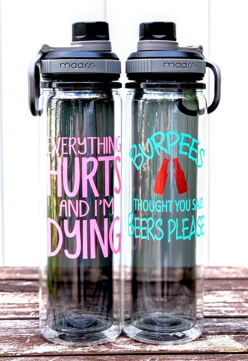 K&ouml;nnte beinhalten: Zwei durchsichtige Wasserflaschen aus Plastik mit schwarzen Deckeln und Tragegriffen. Die linke Flasche tr&auml;gt den Text "Everything Hurts and I'm Dying" in rosa Schrift. Die rechte Flasche tr&auml;gt den Text "Burpees I Thought You Said Beers Please" in t&uuml;rkisfarbener Schrift mit einer roten Grafik von zwei Bierflaschen.