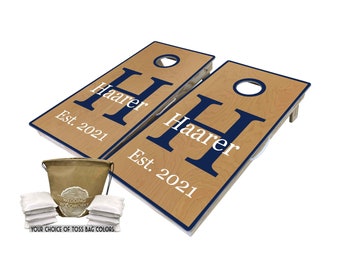 May include: A set of two personalized cornhole boards with a navy blue "H" initial and the text "Haarer Est. 2021" on a light wood background. The set includes a brown canvas bag and eight white bean bags.
