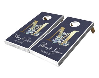 May include: Two navy blue wooden cornhole boards with a gold letter M and floral design. The boards are personalized with the text "Tiffany & Brian" and the date "04.10.2021".