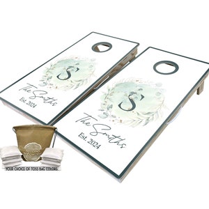 May include: A set of two personalized cornhole boards with a green and white watercolor design featuring a wreath with the letter "S" in the center. The boards are labeled "The Smiths Est. 2024". The set includes a brown canvas bag and eight white bean bags.