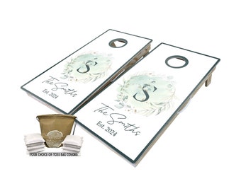 May include: A set of two personalized cornhole boards with a green and white watercolor design featuring a wreath with the letter "S" in the center. The boards are labeled "The Smiths Est. 2024". The set includes a brown canvas bag and eight white bean bags.
