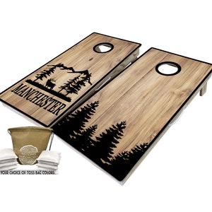 May include: A set of two wooden cornhole boards with a black silhouette of a mountain scene with trees and a deer. The boards are personalized with the name "MANCHESTER". The set includes a brown canvas bag and eight white bean bags.