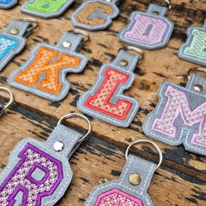 May include: A set of grey felt keychains with embroidered letters in various colours. The letters are in a varsity style font with a white outline. The keychains are arranged in a row on a wooden surface.