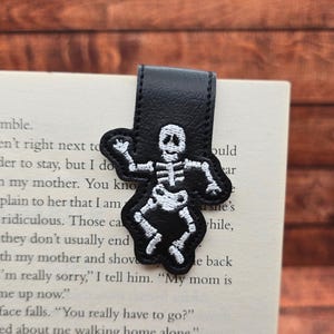 May include: A black magnetic bookmark featuring an embroidered white skeleton figure in a dancing pose. The bookmark is constructed from a textured material and is folded over the top edge of a page containing printed black text on white paper.