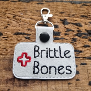 May include: White leather keychain with a red plus sign and the text "Brittle Bones" in black embroidery.
