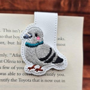 May include: A white leather bookmark featuring an embroidered pigeon design. The pigeon has gray feathers, a teal collar, and pink cheeks. The bookmark is attached to a book with visible text.
