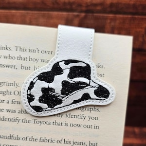 May include: A white leather bookmark featuring a black and white cow print cowboy hat design. The bookmark is attached to a white tab and is inserted into a book.