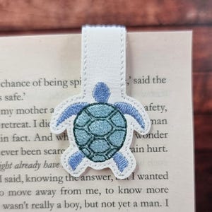 May include: A white magnetic bookmark featuring an embroidered sea turtle with a teal shell and light blue limbs. The turtle is centred on a vertical white strip, designed to clip over the edge of a book page with visible printed text.