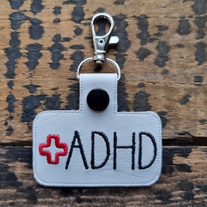 Può includere: Portachiavi in ​​pelle bianca con un segno più rosso e il testo "ADHD" in ricamo nero.
