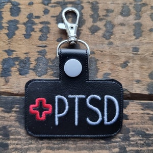 May include: Black leather keychain with a white embroidered cross and the text "PTSD" in white embroidery. The keychain has a silver metal loop.
