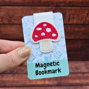 May include: A magnetic bookmark with a red mushroom design and white spots, cream stem, and a white leather strap. The words "Magnetic Bookmark" are printed on a light blue background. The bookmark is designed for use in books.