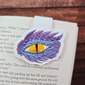 May include: Embroidered bookmark featuring a dragon's eye design. The eye is yellow with black detailing, surrounded by purple and blue stitching. A white leather tab is attached for easy placement in a book. A unique accessory for book lovers.