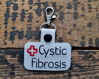 May include: White leather keychain with a red cross and the text "Cystic Fibrosis" in black embroidery. The keychain has a silver metal clip.