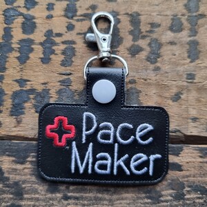 May include: Black rectangular keychain with the words "Pace Maker" embroidered in white, and a red cross symbol. The keychain has a silver clasp and a silver snap closure.