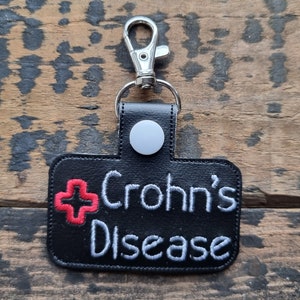 Op de afbeelding: Zwarte leren sleutelhanger met een witte knop en een rood kruis. De tekst "Crohn's Disease" is in wit op de sleutelhanger geborduurd.