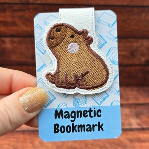 May include: A magnetic bookmark featuring an embroidered brown capybara with a white circular patch on its side. The bookmark is attached to a rectangular card with a light blue background, white book icons, and the text Magnetic Bookmark.