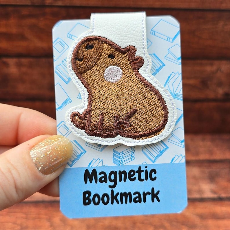 May include: A magnetic bookmark featuring an embroidered brown capybara with a white circular patch on its side. The bookmark is attached to a rectangular card with a light blue background, white book icons, and the text Magnetic Bookmark.