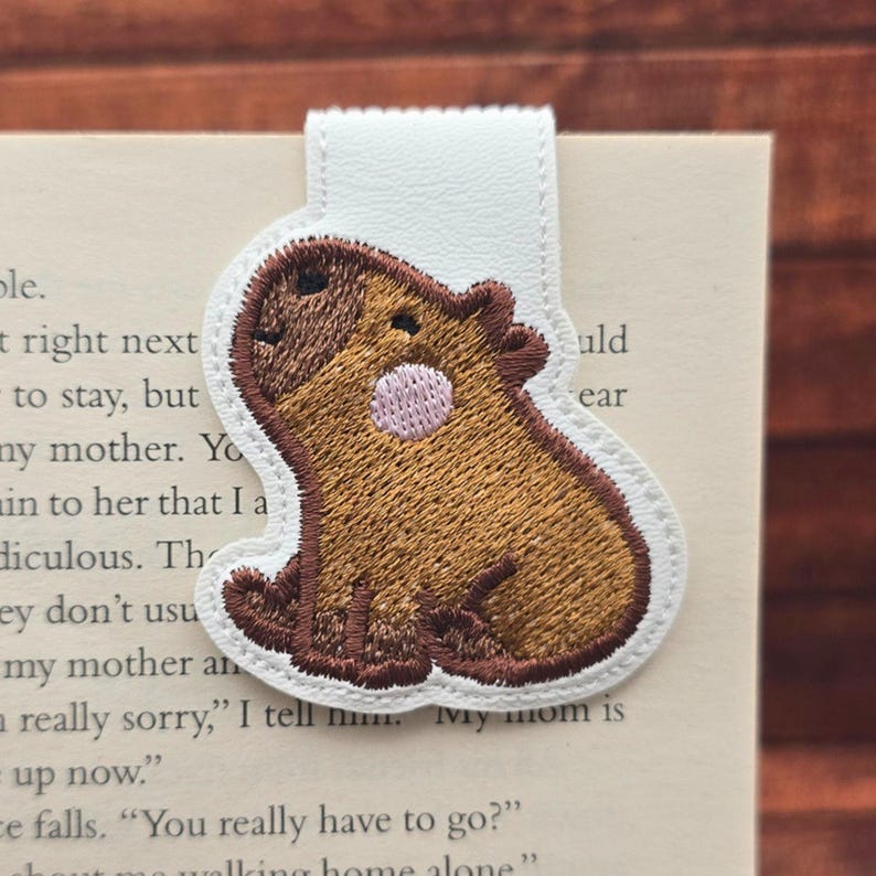 May include: A magnetic bookmark featuring an embroidered capybara design. The capybara is brown with a small pink circular patch on its side and dark brown outlines. It is attached to a white rectangular base that folds over a book page.