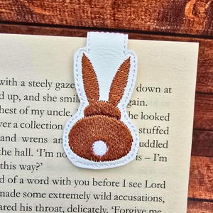 May include: A white bookmark featuring a brown bunny design. The bunny is embroidered with brown thread and has a white tail. The bookmark is on a book with text.