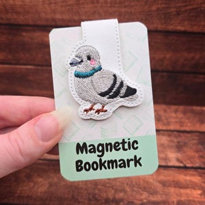 May include: A magnetic bookmark featuring an embroidered gray pigeon with a blue collar and pink cheeks. The bookmark has a white leather strap and a green card with the words "Magnetic Bookmark".