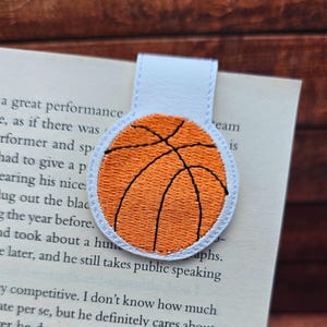 Op de afbeelding: Een boekenlegger met basketbalthema met een oranje basketbalontwerp en zwarte lijnen, geborduurd op een witte achtergrond. De boekenlegger is bevestigd aan een wit lipje.
