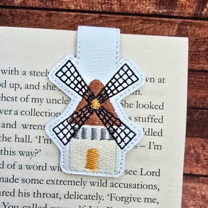 May include: A white windmill-shaped bookmark with black, brown, and yellow embroidery. The bookmark is made of leather and is inserted into the pages of a book. The windmill design features detailed stitching.