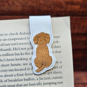 Op de afbeelding: Witte leren boekenlegger met een bruin geborduurd hondenontwerp. De hond wordt van achteren gezien in een zittende positie. De boekenlegger zit in een boek.